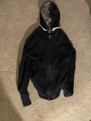 Marinblå hoodie från GANT med dragkedja - Snygg marinblå hoodie från GANT med dragkedja och stor huva. Broderad GANT-logga på bröstet och två stora fickor framtill. Insidan är mjuk och gråmelerad, perfekt för chill dagar. Klassisk collegekänsla och riktigt skön passform.