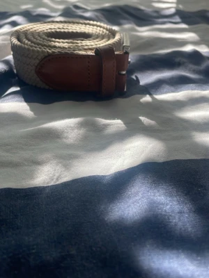 Beige flätat bälte med skinn-detaljer - Snyggt beige flätat bälte med bruna skinn-detaljer vid spännet. Perfekt accessoar för att lyfta din outfit och funkar till både jeans och chinos. Metallspänne i silverfärg ger en stilren touch.
