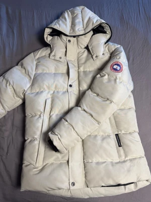Vit dunjacka från Canada Goose - Snygg vit dunjacka från Canada Goose med huva och klassisk logotyp på ärmen. Jackan har dragkedja och knappar framtill, två stora fickor och quiltad design. Perfekt för kalla vinterdagar och riktigt varm tack vare dunfyllningen.