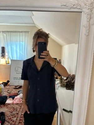 Mörkblå kortärmad skjorta - Stilren mörkblå skjorta med korta ärmar och klassisk krage. Skjortan har knappar framtill och en rak passform som ger en avslappnad look. Perfekt att matcha med både jeans och kjol för en trendig vibe.