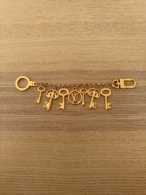  Vuitton nyckelring med guldfärgade berlocker - Säljer en exklusiv nyckelring från Vuitton i guldfärgad metall. Nyckelringen har flera detaljerade nyckelberlocker och LV-logga, samt karbinhake och ring med ingraverad text. Perfekt accessoar för att piffa till väskan eller nyckelknippan.