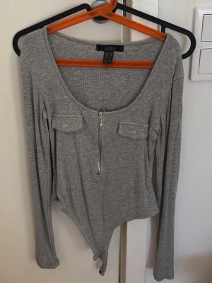 Grå ribbad body med dragkedja - Snygg grå body i ribbat material från VAMPEI. Modellen har lång ärm, djup rundad urringning och dragkedja framtill. Två dekorativa bröstfickor med lock ger en cool detalj. Perfekt att styla till jeans eller kjol.
