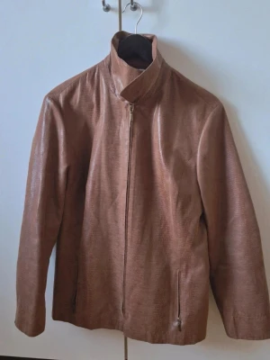 Brun skinnjacka med mönster - Säljer en brun skinnjacka med snyggt reptilmönster och dragkedja framtill. Jackan har två fickor med dragkedja och en klassisk krage. Materialet är glansigt skinn som ger en cool och trendig look.