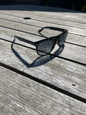 Svarta Ray-Ban solglasögon - Snygga svarta solglasögon från Ray-Ban med klassisk rektangulär form och mörka glas. Bågarna är i plast och har Ray-Ban-loggan på sidan. Perfekta för soliga dagar och ger en cool vibe till din outfit.
