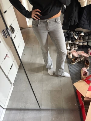 Ljusgrå bootcut jeans - Säljer ett par ljusgrå bootcut jeans med klassisk femficksdesign och låg midja. Jeansen har en snygg, lätt utsvängd passform och är perfekta för dig som gillar en avslappnad men trendig stil. Materialet är denim med en mjuk känsla.