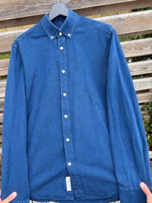 Blå skjorta från Tiger of Sweden - Snygg blå skjorta från Tiger of Sweden i slim fit. Skjortan har klassisk krage, vita knappar framtill och långa ärmar. Tillverkad i ett mjukt bomullstyg som känns skönt mot huden. Perfekt för dig som gillar stilrena och tidlösa plagg.
