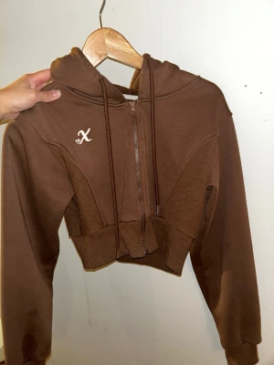 Brun croppad hoodie med dragkedja - Säljer en brun croppad hoodie med dragkedja framtill och huva. Hoodien har ett broderat vitt X på bröstet och ribbade detaljer vid midjan. Tillverkad i mjukt sweatshirtmaterial och har långa ärmar. Perfekt för en avslappnad och trendig look.