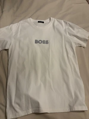 Vit BOSS t-shirt med tryck - Snygg vit t-shirt från BOSS med klassiskt rund halsringning och ett stilrent BOSS-tryck i svart och vitt på bröstet. Tillverkad i mjuk bomull som känns skön mot huden. Perfekt för dig som gillar enkel och clean stil.