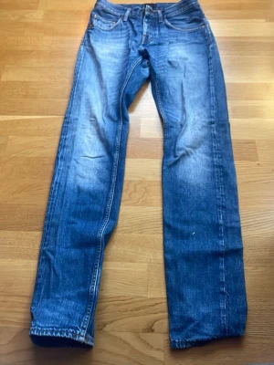 Blå raka jeans med slitningar - Säljer ett par klassiska blå jeans med raka ben och snygga slitningar framtill. Jeansen har fem fickor, bälteshällor och kontrastsömmar. Materialet är denim i bomull och passformen är relaxed fit. Perfekta till en avslappnad stil.