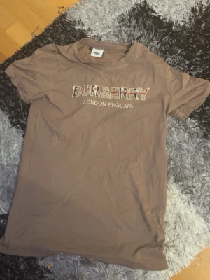Brun Burberry t-shirt med logga - Säljer en brun t-shirt från Burberry med klassisk logga och texten 'London England' tryckt på bröstet. T-shirten har rund hals och korta ärmar. Materialet känns mjukt och skönt, perfekt till en avslappnad stil.