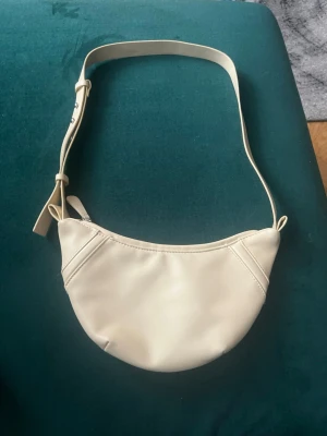 Krämvit axelväska i halvmåneform - Snygg axelväska i krämvitt konstläder med halvmåneformad design och justerbart axelband. Väskan har minimalistiska sömdetaljer och en enkel dragkedja upptill. Man kan göra den till olika storlekar. Helt ny väska som inte alls har använts. Det finns mycket plats i väskan. 