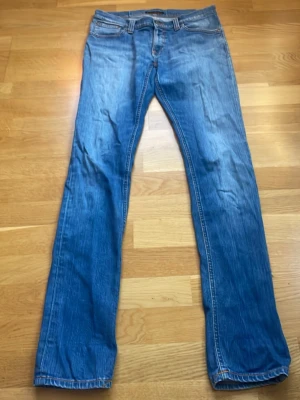 Blå raka jeans från Nudie Jeans - Snygga blå jeans från Nudie Jeans med rak passform och klassisk femficksdesign. Jeansen har orangea sömmar och är tillverkade i denim av bomull. Perfekta för en avslappnad och stilren look.