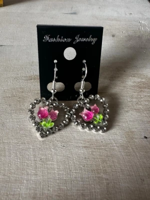 Hjärformade örhängen med blommor - Säljer ett par hjärtformade örhängen dekorerade med glittrande stenar runt kanten och rosa blommor med gröna blad i mitten. Örhängena har krokar i silverfärg och ger en söt och romantisk look. Perfekta för dig som gillar detaljer och blommigt.