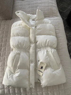 Vit dunväst med hög krage - Snygg vit dunväst från POLOMOOD med hög krage och breda vadderade paneler. Västen har två fickor framtill och elastisk midja för en schysst passform. Stängs med dragkedja och tryckknappar. Perfekt för lager på lager under kyliga dagar.