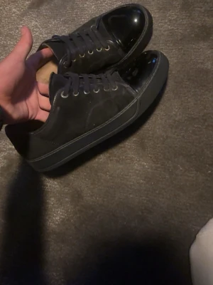 Lanvin skor - Snygga svarta sneakers från Lanvin med klassisk låg profil. Skorna har en unik lackad tå och ovandel i mocka och läder, samt svarta skosnören och sula. Perfekta för dig som gillar stilrena och exklusiva sneakers med en twist. Det är en liten repa på vänstra skon men inget annat än det. Skorna är uk 6