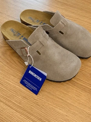 Beige birkenstock boston tofflor - Säljer ett par snygga beige tofflor från Birkenstock helt nya. De har en justerbar rem med metallspänne och präglad logga på sidan. Fotbädden är ergonomisk och sulan är i svart gummi för extra komfort. Pris kan diskuteras vid snabb affär!