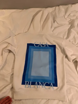 Vit hoodie med blått tryck från Casablanca - Säljer en vit hoodie från Casablanca med stor grafisk blå rektangel på ryggen och texten 'CASA BLANCA'. På bröstet finns en mindre blå ruta. Hoodien har huva med dragsko och magficka. Perfekt för dig som gillar stilrena men ändå unika plagg.