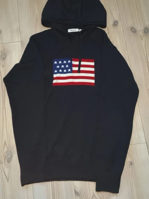 Svart hoodie med USA-flagga från RYVLS - Svart hoodie från RYVLS med stor amerikansk flagga på bröstet. Klassisk huva med snörning och långärmad modell. Perfekt för dig som gillar streetwear och vill sticka ut lite extra.