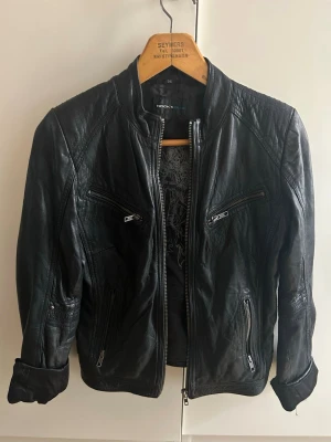 Svart skinnjacka med dragkedjor - Snygg svart skinnjacka i klassisk bikerstil med flera dragkedjeförsedda fickor framtill och detaljerade sömmar. Jackan har ståkrage och justerbara spännen i midjan. Perfekt för dig som vill ha en cool och tidlös look.