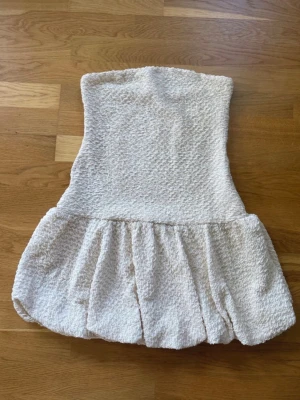 Krämvit axelbandslös kort klänning H&M - Superfin krämvit axelbandslös kort klänning från H&M med bubblig struktur och volangig kjol. Tillverkad i återvunnen polyester och elastan för en bekväm och stretchig passform. Perfekt för dig som gillar trendiga och unika plagg.