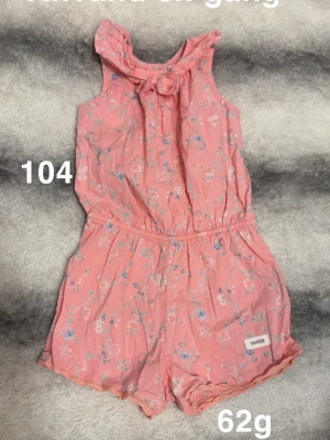 Korallfärgad blommig jumpsuit med rosett - Söt korallfärgad jumpsuit med blommönster och en dekorativ rosett vid halsen. Ärmlös modell med resår i midjan och korta ben. Perfekt för varma dagar och enkel att matcha med sneakers eller sandaler.🌸 Använd en gång. Storlek 104