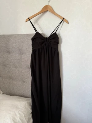 Svart långklänning med smala axelband - Svart långklänning med smala justerbara axelband och rynkad detalj vid bysten. Klänningen har en enkel och stilren design, perfekt för dig som gillar minimalistiskt mode. Tillverkad i ett mjukt och lätt material som faller snyggt.