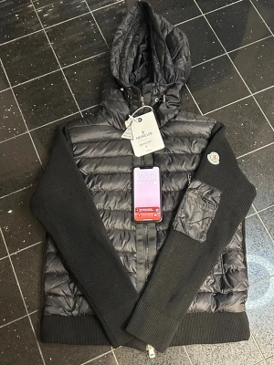 Svart cardigan från Moncler - Säljer en svart pufferjacka från Moncler med stickade ärmar och huva. Jackan har dragkedja framtill, ficka på ena ärmen och klassisk Moncler-logga på axeln. Materialet är en mix av glansigt nylon och ribbstickad bomull vilket ger en snygg kontrast.