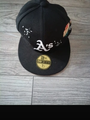 Svart New Era 59FIFTY A's keps - Säljer en svart New Era 59FIFTY keps med A's-logga framtill och broderade blommor på sidan. Kepsen har platt skärm och klassisk snapback-look. Materialet är troligtvis bomull eller polyester. Perfekt för dig som gillar streetwear och sportig stil.