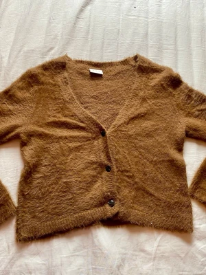 Brun fluffig kofta från Vila - Mysig brun kofta från Vila med fluffig textur och tre knappar framtill. Koftan har v-ringning och långa ärmar, perfekt för lager på lager och en chill look. Passar dig som gillar mjuka material och vill ha en trendig, avslappnad stil.