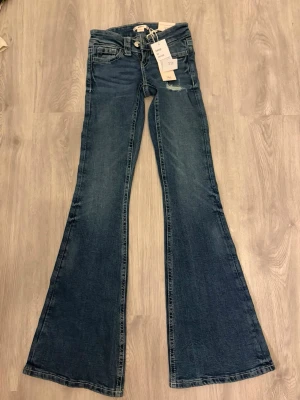 Blå bootcut jeans med broderade fickor - Säljer ett par blå bootcut jeans med snygg slitning och broderade detaljer på bakfickorna. Jeansen har låg midja, klassisk femficksmodell och vida ben. Perfekta för dig som gillar retrostil och vill sticka ut lite extra.