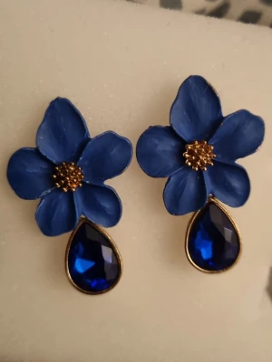 Blå blomörhängen med droppformad sten - Unika örhängen med stora blå blommor i plast och guldfärgade detaljer i mitten. Nedanför hänger en droppformad, djupblå glittrande sten infattad i guldfärgad metall. Perfekta för dig som vill sticka ut med färg och form.