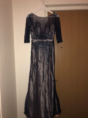 Mörkblå långklänning med paljetter - Elegant mörkblå långklänning med transparent mesh och vackra paljettbroderier. Klänningen har trekvartsärm och markerad midja med dekorativt band. Perfekt för speciella tillfällen där du vill sticka ut.