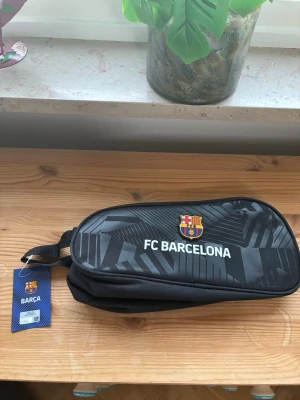 Svart FC Barcelona necessär - Snygg svart necessär med FC Barcelona-logga och texten 'FC BARCELONA' på framsidan. Väskan har dragkedja, bärhandtag och sportig design med mönstrade detaljer. Perfekt för att förvara småsaker eller hygienartiklar. Materialet är slitstark textil.