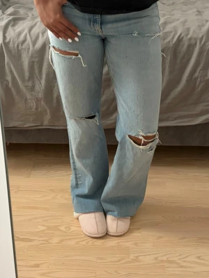 Zara baggy jeans - Säljer ett par ljusblå jeans med raka ben och stora slitningar vid knäna och låret. Jeansen har en avslappnad passform och klassisk femficksdesign. Perfekta för en trendig och avslappnad stil.