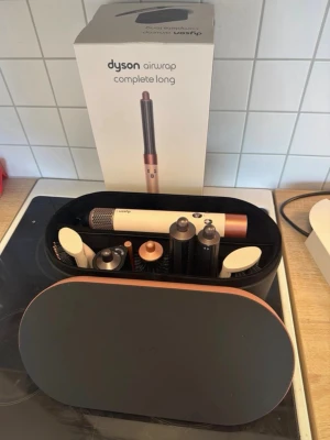 Dyson Airwrap Complete Long hårstyler - Säljer en Dyson Airwrap Complete Long i elegant vit och kopparfärg. Setet innehåller flera stylingtillbehör som borstar och locktillbehör, allt i ett snyggt svart och kopparfärgat etui. Perfekt för att skapa olika frisyrer med avancerad teknik.
