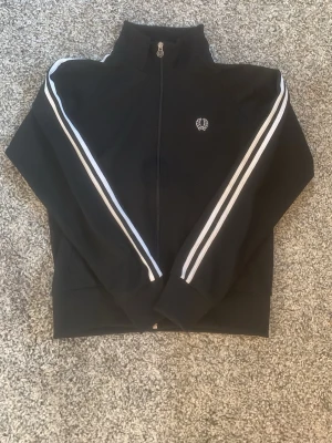 Svart Fred Perry ziptröja med ränder - Snygg svart ziptröja från Fred Perry med vita dubbla ränder längs ärmarna och broderad logga på bröstet. Hög krage och hel dragkedja framtill. Perfekt för en sportig och stilren look.