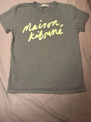 Grön Maison Kitsuné t-shirt - Snygg grön t-shirt från Maison Kitsuné med gul text framtill. Klassisk rund halsringning och korta ärmar. Tillverkad i mjuk bomull som känns skön mot huden. Perfekt för dig som gillar stilrena plagg med en twist.