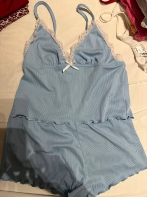 Ljusblått ribbat pyjamasset - Sött ljusblått pyjamasset i ribbat material med spetsdetaljer i vitt vid bysten och tunna axelband. Setet har en liten rosett framtill och shorts med vågig kant. Perfekt för dig som gillar mjuka och bekväma underkläder.