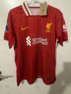 Liverpool matchtröja röd herr - Snygg röd Liverpool matchtröja med gula och vita detaljer, klubbmärke och Nike-logga på bröstet. Sponsortryck på framsidan och ärmarna. Namnet 'Gravenberch' och nummer 38 på ryggen. Tillverkad i lätt och ventilerande material, perfekt för fotboll eller träning. 