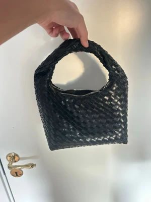 Svart flätad handväska - Snygg svart handväska med flätad design och kort handtag. Väskan har en mjuk, fyrkantig form och stängs med dragkedja. Perfekt accessoar för dig som gillar stilrena detaljer och trendig look.