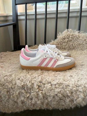Adidas Samba rosa och vita sneakers - Säljer ett par Adidas Samba sneakers i vitt och ljusrosa med klassiska tre ränder på sidan och gummisula. Skorna har mockadetaljer vid tån och är låga i modellen. Perfekta för dig som vill ha en sportig och trendig look. Använda vid 1 tillfälle.