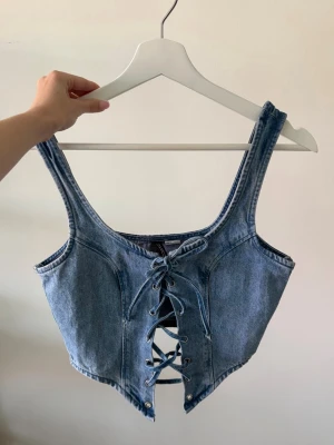 Blå denim korsettopp med snörning - Snygg blå korsettopp i denim med snörning framtill. Toppen har en croppad passform och markerad midja.