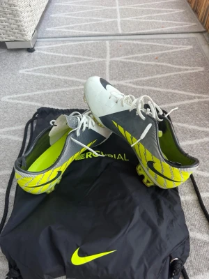 Nike Mercurial vita och gula fotbollsskor - Säljer ett par Nike Mercurial fotbollsskor i vitt, svart och neongult. Skorna har snörning, låg profil och en lätt ovandel i syntetmaterial. Sulan har dobbar för bra grepp på planen. Kommer med en svart Mercurial-påse med neongul logga.