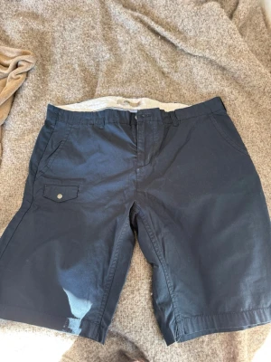 Mörkblåa shorts från 8848 Altitude - Snygga mörkblåa shorts från 8848 Altitude med klassisk passform. De har fickor fram, en ficka med lock och knapp på sidan samt bälteshällor. Materialet är bomullstyg som känns slitstarkt och bekvämt. Perfekta för sommardagar!