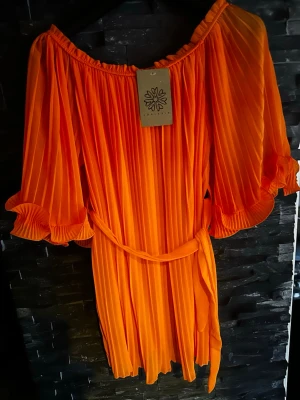 orange plisserad blus - Säljer en , orange blus  med plisserat tyg och volangdetaljer på ärmarna. Blusen har offshoulder-modell, trekvartsärm och ett matchande midjeband för snygg passform. 