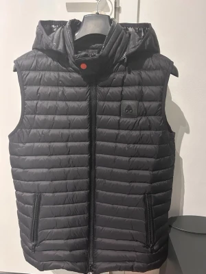 Svart dunväst från Moose Knuckles - Snygg svart herr dunväst från Moose Knuckles med huva och dragkedja framtill. Västen har två fickor med dragkedja och diskret logga på bröstet. Quiltad design i lätt och smidigt material som håller värmen utan att bli klumpig.