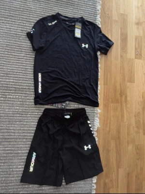 Sett Under Armour - Säljer ett svart träningsset från Under Armour med t-shirt och shorts. Setet har logga och textdetaljer i vitt och reflekterande färg. Tillverkat i ett lätt och snabbtorkande material, perfekt för träning eller sportaktiviteter.