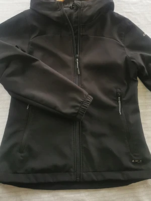 Softshell jacka 152 - Svart softshell från Icepeak med huva och dragkedja framtill. Jackan har två sidofickor med dragkedja, elastiska muddar och slätt yttertyg. Insidan är fodrad med mjuk fleece för extra komfort. Perfekt för blåsiga dagar och enkel att matcha.