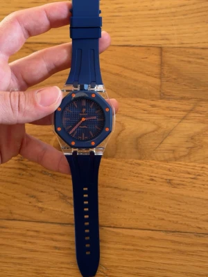 Blå sportig klocka med orange detaljer - Snygg klocka med marinblått silikonarmband och matchande blå urtavla. Urtavlan har orange markeringar och visare som ger en cool kontrast. Boetten är transparent vilket ger en modern känsla. Perfekt accessoar för dig som gillar färg och stil.