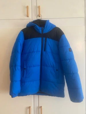 Blå pufferjacka från BOSS - Snygg blå pufferjacka från BOSS med svarta detaljer över axlarna och svart dragkedja. Jackan har hög krage, bröstficka med dragkedja och två sidofickor. BOSS-logga på vänster ärm. Perfekt för kalla vinterdagar.